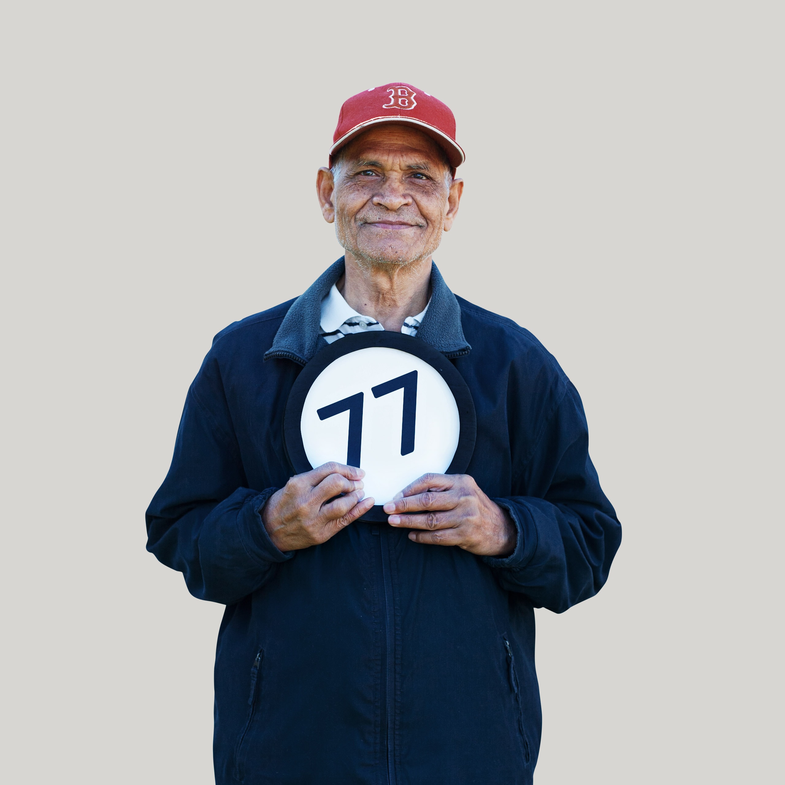 77 year old man wears a baseball cap and a closed mouth smile. He looks directly into the lens  while holding to his chesy a token with the number 77 on it. This digital image is part of the 1 to Infinity portrait photography series by Danny Goldfield.