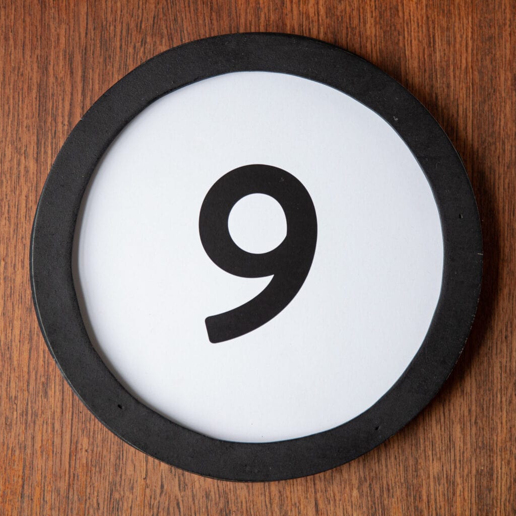 A circle shaped token fills the square image. The token has a 1 inch black circular frame around it. In the center is a black number on a white background. The token lies on a teak wood table. This digital image is part of the 1 to Infinity portrait photography series by Danny Goldfield.