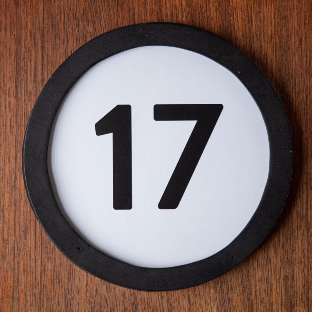 A circle shaped token fills the square image. The token has a 1 inch black circular frame around it. In the center is a black number on a white background. The token lies on a teak wood table. This digital image is part of the 1 to Infinity portrait photography series by Danny Goldfield.