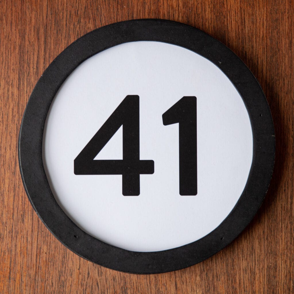 A circle shaped token fills the square image. The token has a 1 inch black circular frame around it. In the center is a black number on a white background. The token lies on a teak wood table. This digital image is part of the 1 to Infinity portrait photography series by Danny Goldfield.