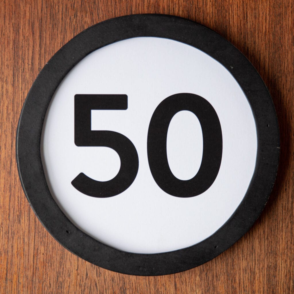 A circle shaped token fills the square image. The token has a 1 inch black circular frame around it. In the center is a black number on a white background. The token lies on a teak wood table. This digital image is part of the 1 to Infinity portrait photography series by Danny Goldfield.