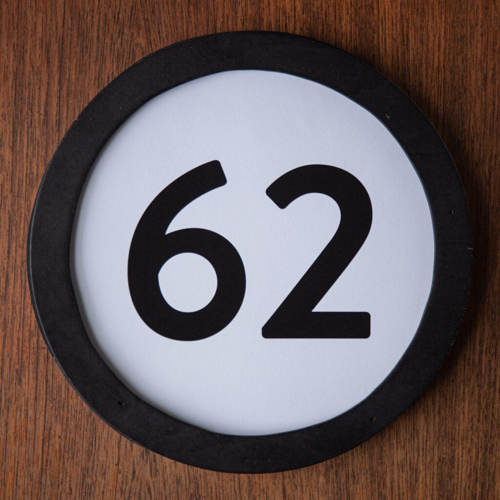 A circle shaped token fills the square image. The token has a 1 inch black circular frame around it. In the center is a black number on a white background. The token lies on a teak wood table. This digital image is part of the 1 to Infinity portrait photography series by Danny Goldfield.