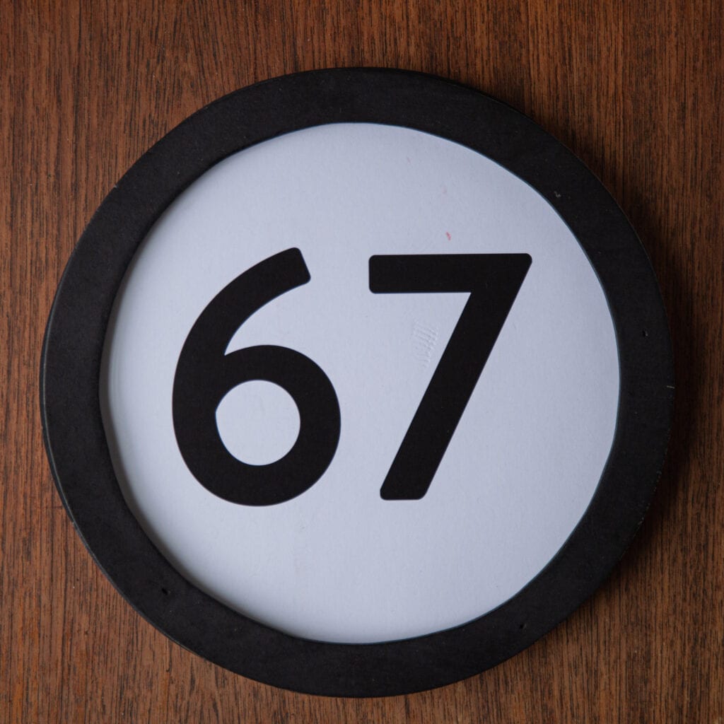 A circle shaped token fills the square image. The token has a 1 inch black circular frame around it. In the center is a black number on a white background. The token lies on a teak wood table. This digital image is part of the 1 to Infinity portrait photography series by Danny Goldfield.