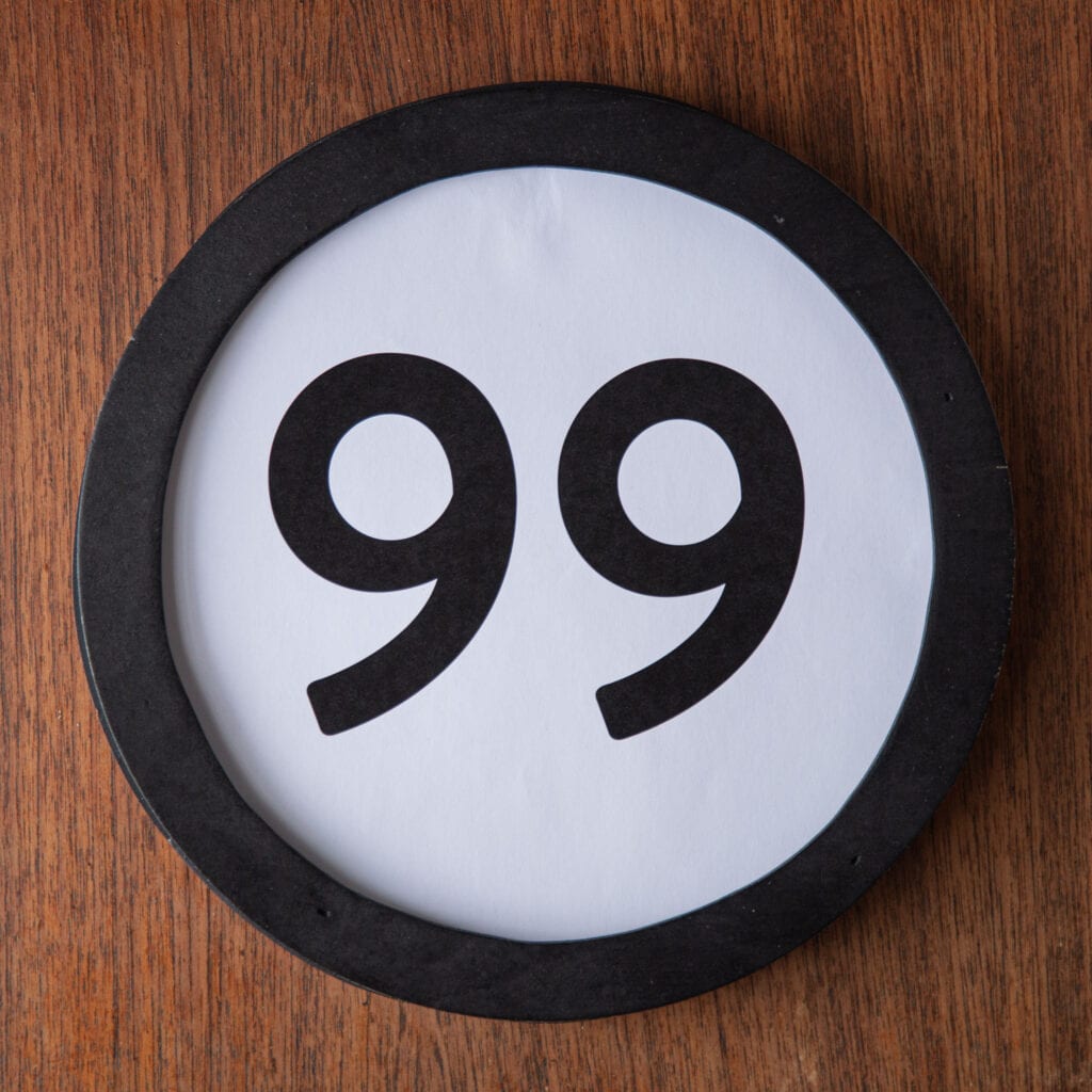 A circle shaped token fills the square image. The token has a 1 inch black circular frame around it. In the center is a black number on a white background. The token lies on a teak wood table. This digital image is part of the 1 to Infinity portrait photography series by Danny Goldfield.