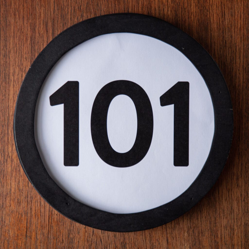 A circle shaped token fills the square image. The token has a 1 inch black circular frame around it. In the center is a black number on a white background. The token lies on a teak wood table. This digital image is part of the 1 to Infinity portrait photography series by Danny Goldfield.